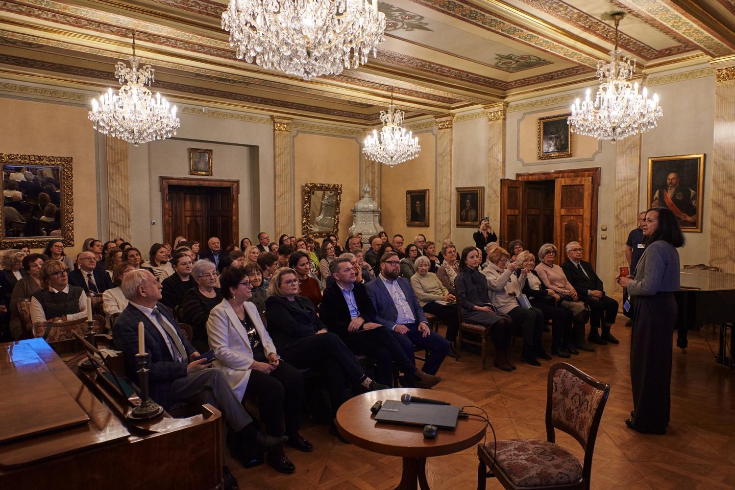 W eleganckiej, zabytkowej sali pałacowej odbywa się wykład o muzeoterapii. Prelegentka stoi przy stole z laptopem i mikrofonem, zwracając się do licznej publiczności, która uważnie słucha, robi notatki i fotografuje. Kryształowe żyrandole, historyczne portrety i bogate zdobienia tworzą atmosferę intelektualnej wymiany myśli w otoczeniu dziedzictwa kulturowego.