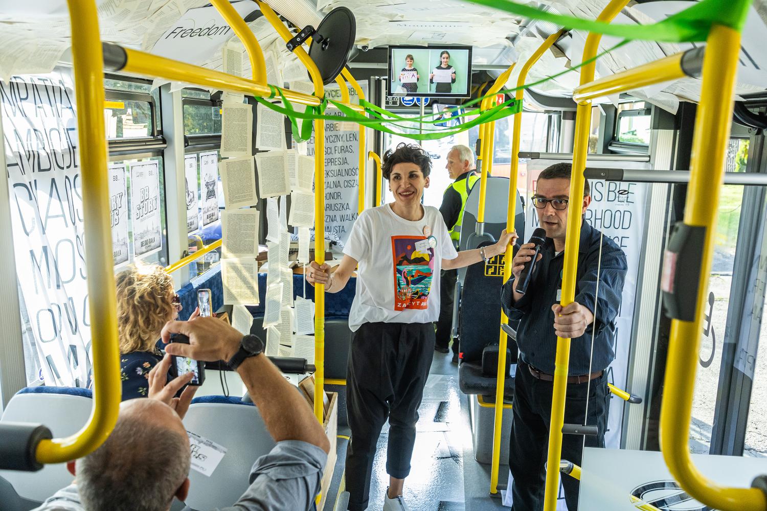 Wnętrze miejskiego autobusu zamienionego w przestrzeń artystyczną. Na poręczach zawieszono kartki z tekstami, zielone wstążki, a ściany ozdobiono czarno-białymi plakatami z hasłami. Na pierwszym planie kobieta w białym T-shircie z kolorowym nadrukiem i uśmiechem trzyma się poręczy. Obok niej mężczyzna z mikrofonem przemawia do publiczności. W tle osoby fotografujące i kierowca w kamizelce odblaskowej.