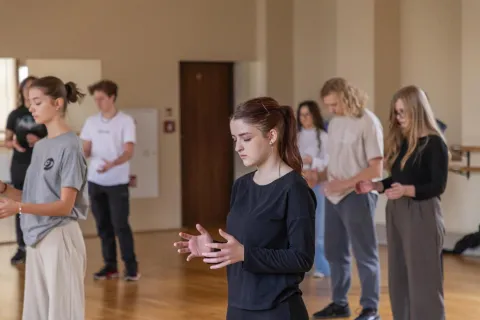 Na zdjęciu uczestnicy warsztatów prowadzonych przez **Karola Pruciaka** koncentrują się na wykonywanych ćwiczeniach. Grupa młodych ludzi, w luźnych, wygodnych strojach, stoi w przestronnej sali z drewnianą podłogą. Wszyscy mają zamknięte oczy lub są głęboko skupieni, wykonując subtelne ruchy dłońmi, co sugeruje praktykę opartą na uważności i świadomości ciała.