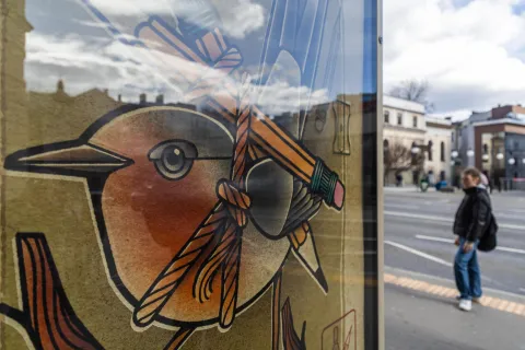 Zbliżenie ilustracji ptaka oplecionego ołówkami, widocznej na przystankowym plakacie. W tle, przez szybę, odbijają się chmury i zabudowa miejska, a po chodniku przechodzi kobieta z plecakiem.