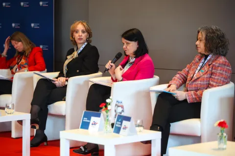 Panel dyskusyjny z udziałem czterech kobiet siedzących na białych fotelach; jedna z panelistek mówi do mikrofonu, jest to wiceministra zdrowia Katarzyna Maria Kacperczak. Na stoliku przed nimi stoją tabliczki z nazwiskami, szklanki wody i małe wazoniki z kwiatami.