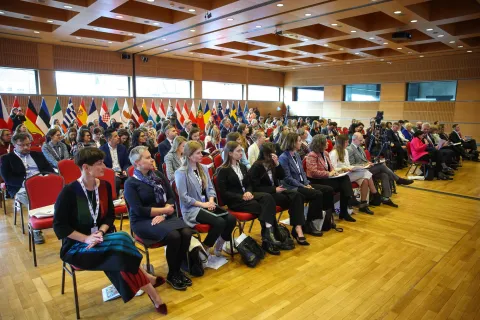 „Widok na publiczność podczas konferencji międzynarodowej – uczestnicy siedzą na czerwonych krzesłach w sali konferencyjnej, w tle rząd flag państw europejskich.”