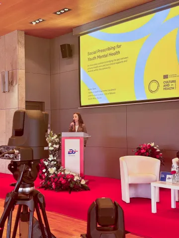 Edyta Ruta z Instytutu Kultury Miejskiej – Miasta Splotów przemawia podczas konferencji, stojąc przy mównicy z logo POLAND25.EU. Na ekranie za nią widnieje slajd prezentacji zatytułowany ‘Social Prescribing for Youth Mental Health’. Wokół mównicy ustawiono kompozycje kwiatowe i sprzęt nagrywający.