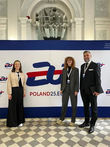 Na zdjęciu widzimy trzy osoby stojące na tle ścianki z logo „POLAND25.EU”. Ścianka znajduje się wewnątrz budynku o klasycystycznej architekturze – w tle widać łukowate sklepienie, kolumny oraz rzeźbiony napis „INFO TICKETS”. Podłoga wyłożona jest biało-szarą szachownicą. Wszyscy uczestnicy mają identyfikatory, sugerujące udział w oficjalnym wydarzeniu.