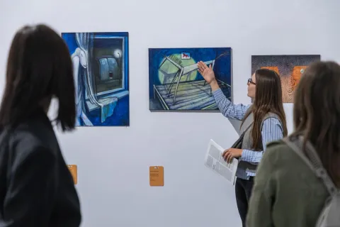 Na zdjęciu przewodniczka oprowadza grupę osób po wystawie sztuki współczesnej. Wskazuje na jedno z ekspresyjnych, surrealistycznych obrazów o intensywnych niebieskich barwach, trzymając w ręku katalog wystawy. Uczestnicy z uwagą słuchają, analizując prezentowane dzieła. Atmosfera galerii jest kameralna i sprzyja refleksji nad sztuką.