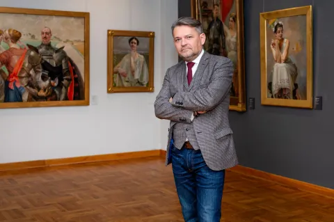 Na zdjęciu elegancko ubrany mężczyzna stoi z założonymi rękami w sali muzealnej, otoczony klasycznymi obrazami w ozdobnych ramach. Jego pewna siebie postawa i bezpośrednie spojrzenie sugerują zaangażowanie w sztukę i kulturę. Wnętrze z drewnianą podłogą i stonowanym oświetleniem tworzy atmosferę dostojności i artystycznej refleksji.